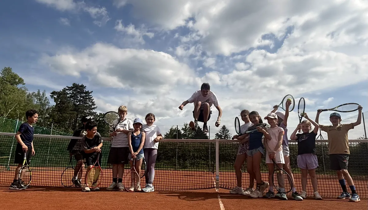 Eine Gruppe Kinder springt gleichzeitig mit erhobenen Schlägern auf dem Sandplatz in die Luft, darüber ein weiter Wolkenhimmel