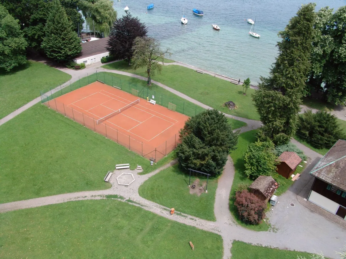 Luftaufnahme des Seeplatzes im Navillegut: ein Sandplatz zwischen Wiese und alten Bäumen, dahinter der Zürichsee mit ankernden Booten
