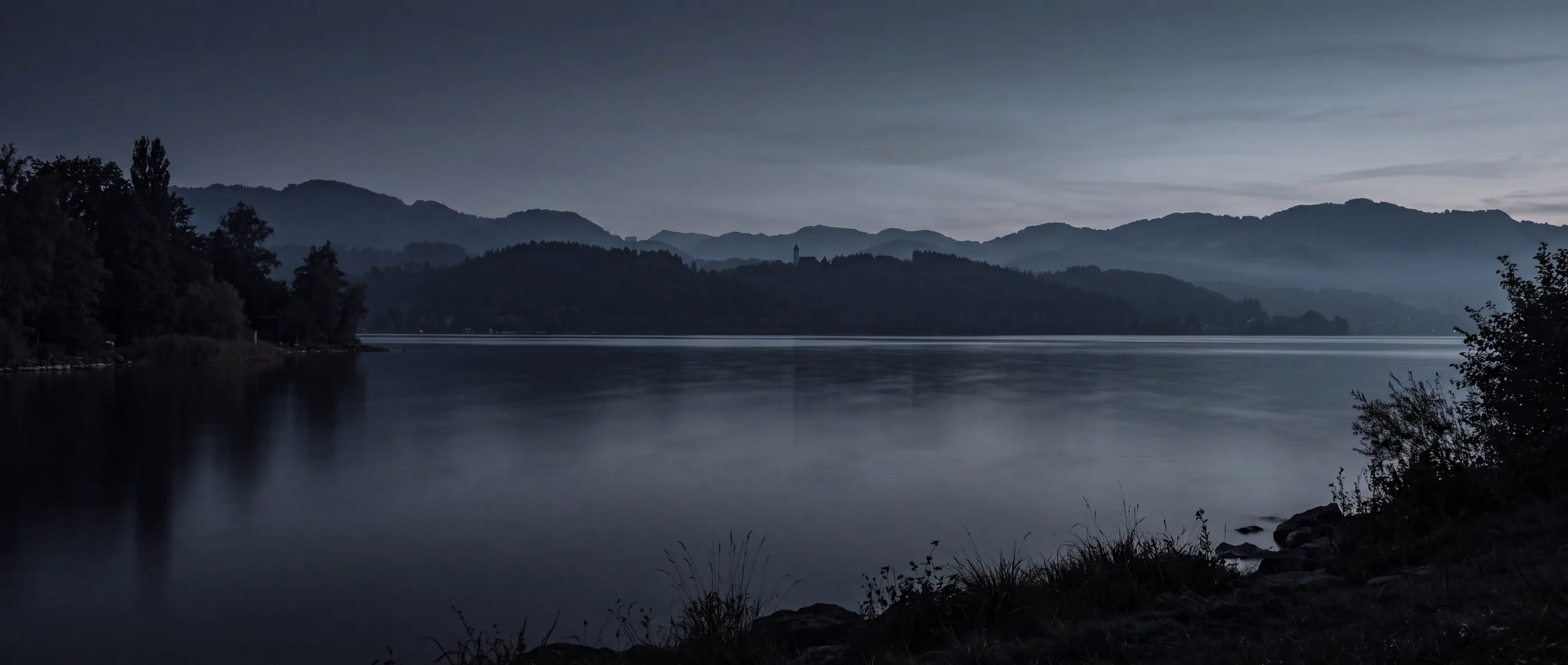 Der Zürichsee bei Kilchberg zur blauen Stunde: gestaffelte Hügelzüge im Dunst, ruhiges Wasser, ein Kirchturm auf dem Grat